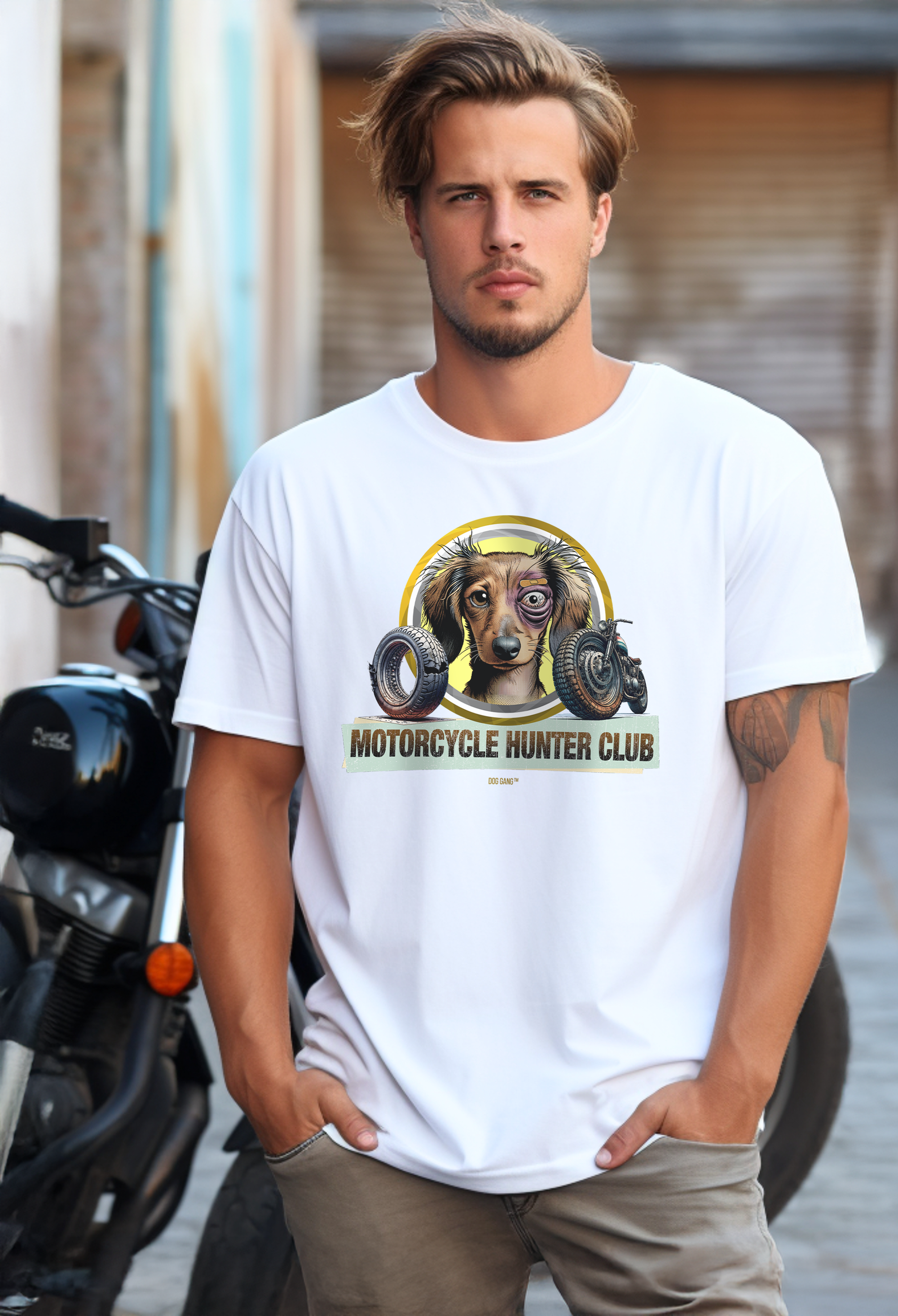 Ein Mann mit kurzen Haaren steht auf einer Straße vor einem Motorrad. Er trägt ein weißes T-Shirt von Dog Gang mit einem Motiv des Motorcycle Hunter Club, das ein leicht zersaustes aber glückliches Hundegesicht mit einem blauen Auge und angebissene Motorradteile zeigt.